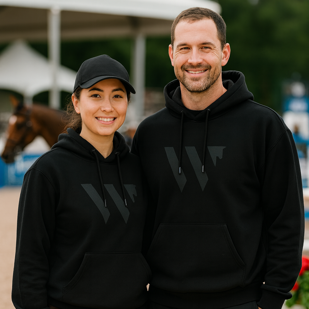 Two people wearing black hoodies with a logo, standing in an outdoor equestrian setting.